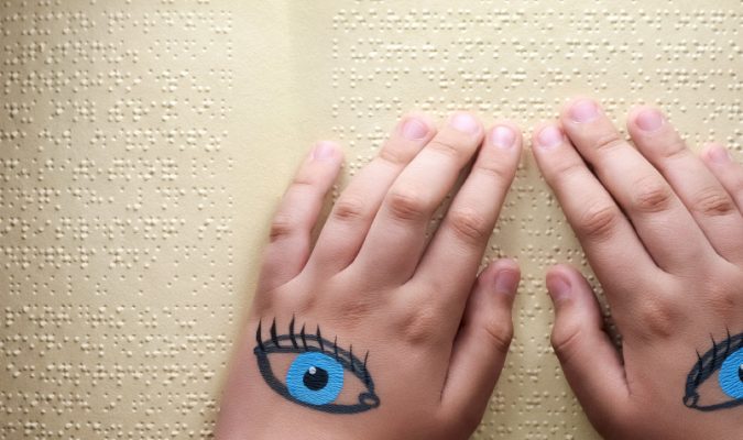 Fotografia di mani su foglio braille. Sul dorso delle mani sono disegnati due occhi.