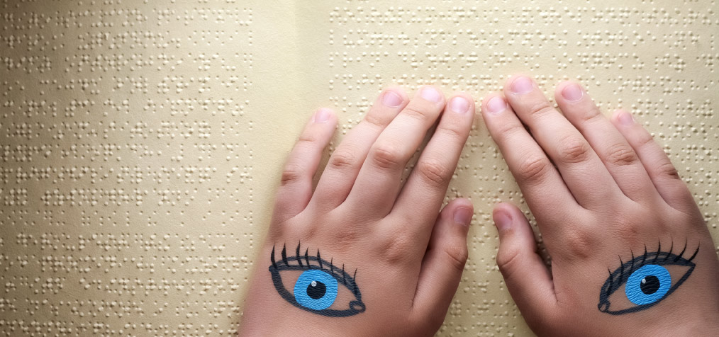 Fotografia di mani su foglio braille. Sul dorso delle mani sono disegnati due occhi.