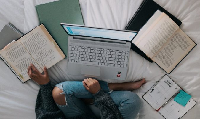 Fotografia di una ragazza seduta sul letto davanti a un laptop e circondata da libri